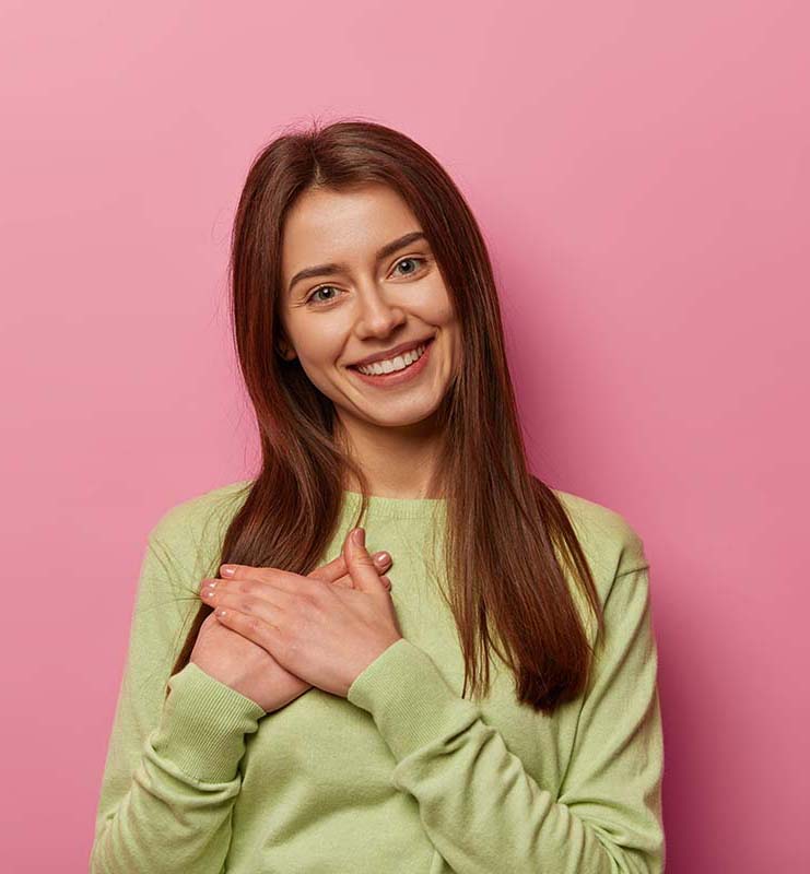 Adorable European woman pressed hands to palm, sees heartwarming scene, looks with admiration and gratitude, smiles gently, wears green jumper, isolated on pink wall copy space area thanks for present